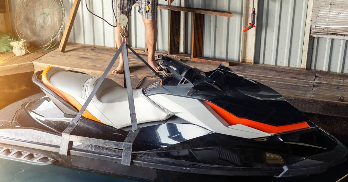 A man is standing on a wooden dock and using a handheld device to lift a black and white jet ski out of the water.