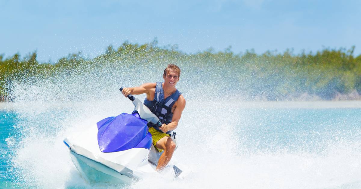 A man wearing a blue life jacket while riding a blue and white jet ski on a sunny day. The water is splashing around him.