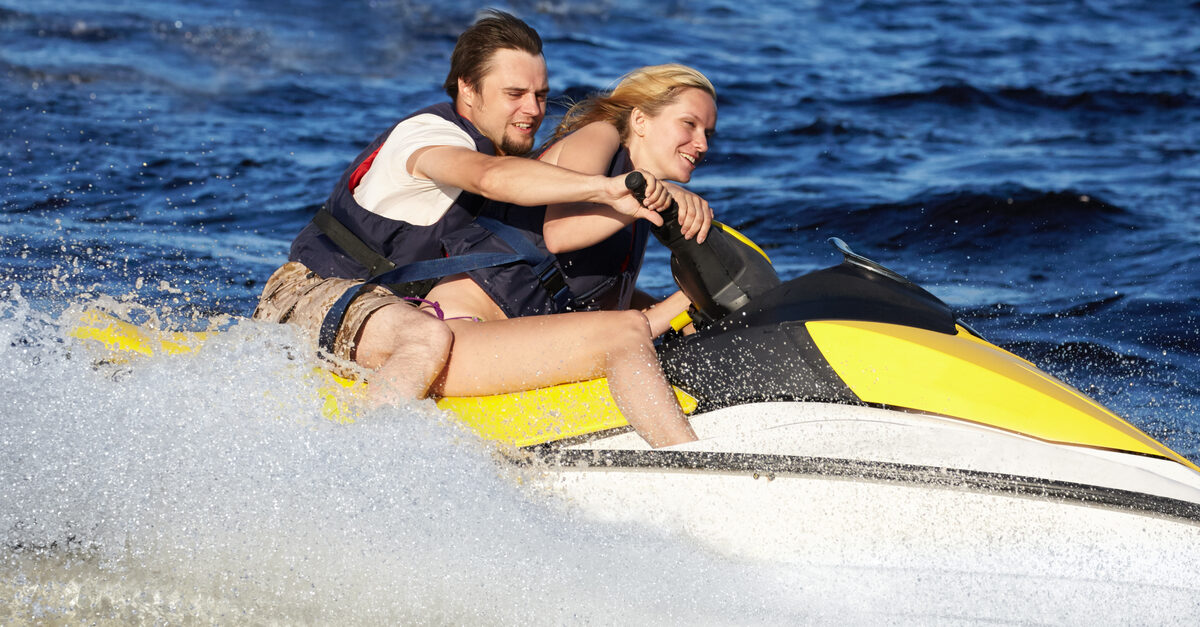 A man and a woman wearing life jackets while riding a yellow and white jet ski. The water is splashing up around them.