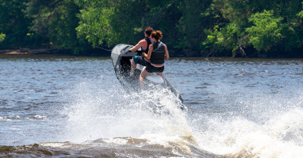 A man and a woman riding on a jet ski on a sunny day. The jet ski is airborne and making waves around them.