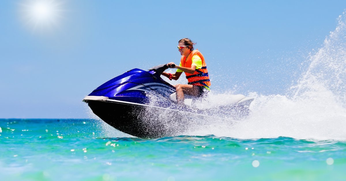 A young man in an orange life jacket and reflective sunglasses smiling while riding a blue jet ski on a sunny day.