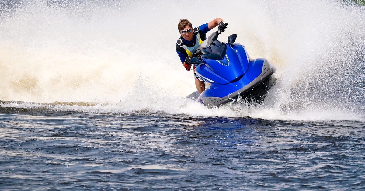 A man wearing sunglasses making a sharp turn on a blue jet ski while waves splash around the watercraft.