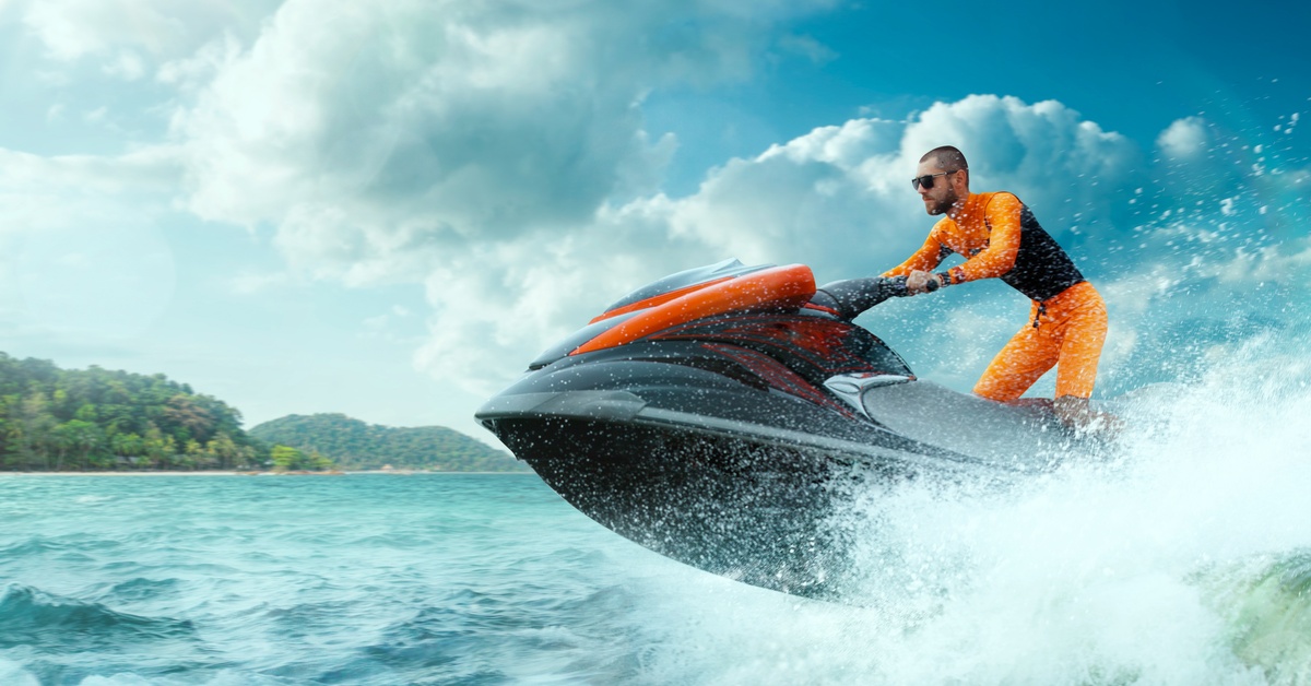 A man in an orange-and-black outfit riding a personal watercraft. It is making waves as the front end glides above the water.