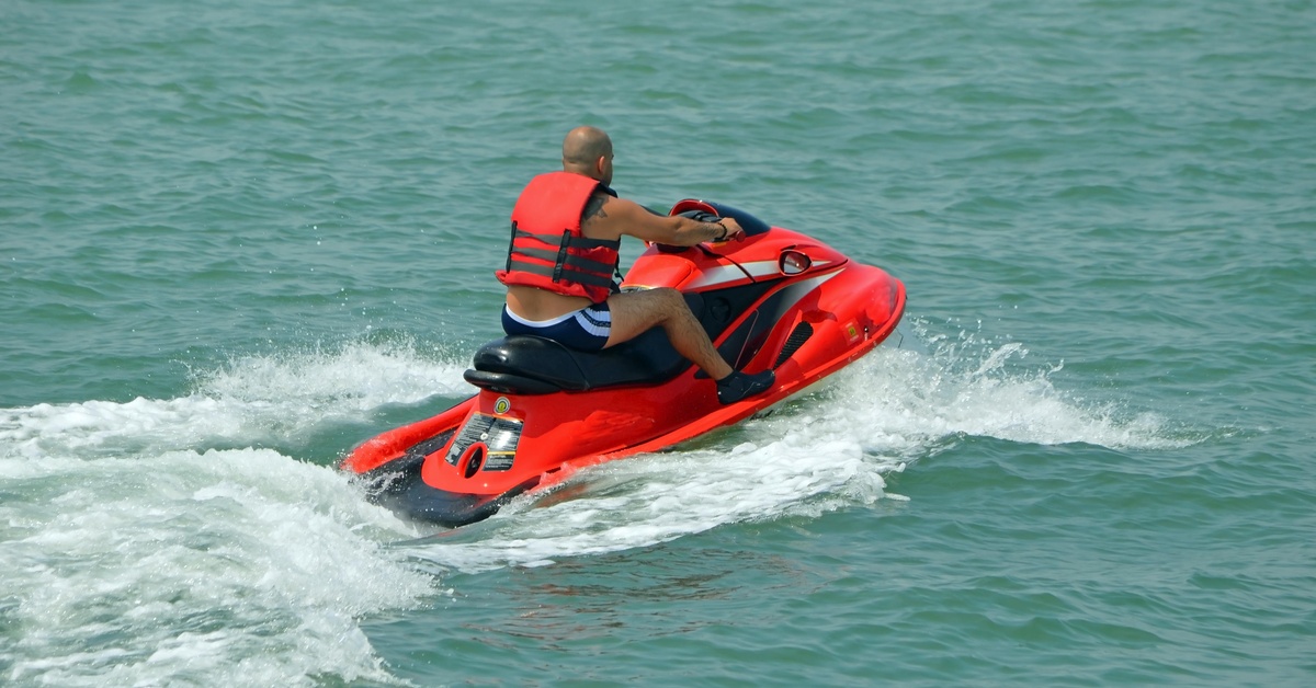 Rearview of a man in an life jacket and blue swim trunks riding an orange jet ski in a large body of water.