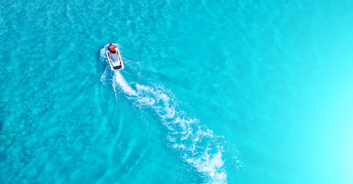 An aerial view of a person riding a jet ski on a large body of water. The water is clear and bright blue.
