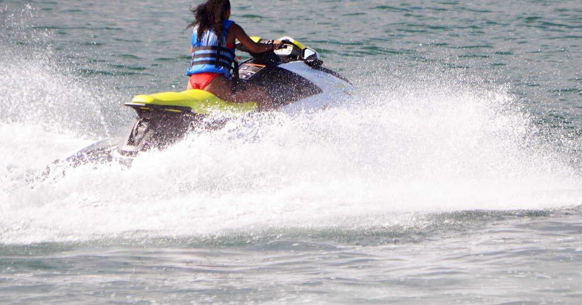 A woman in a blue life jacket rides a jet ski, splashing water up behind her, on a beautiful sunny day.