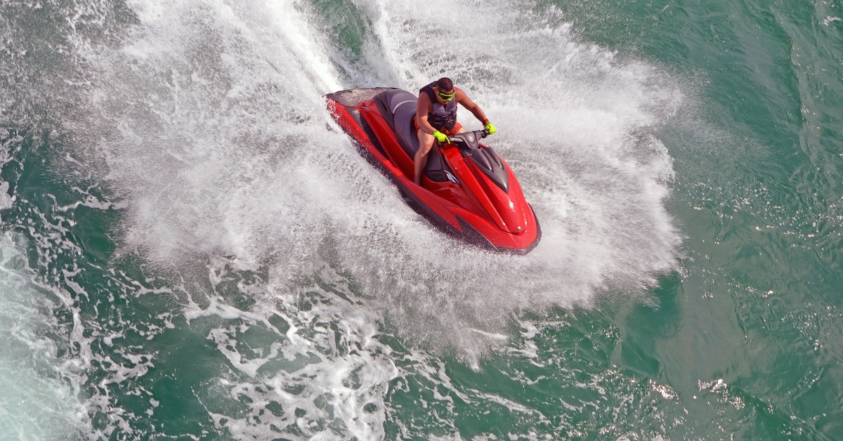 A man rides a red jet ski over the water, pushing white water around him. He's wearing a life vest, gloves, and goggles.