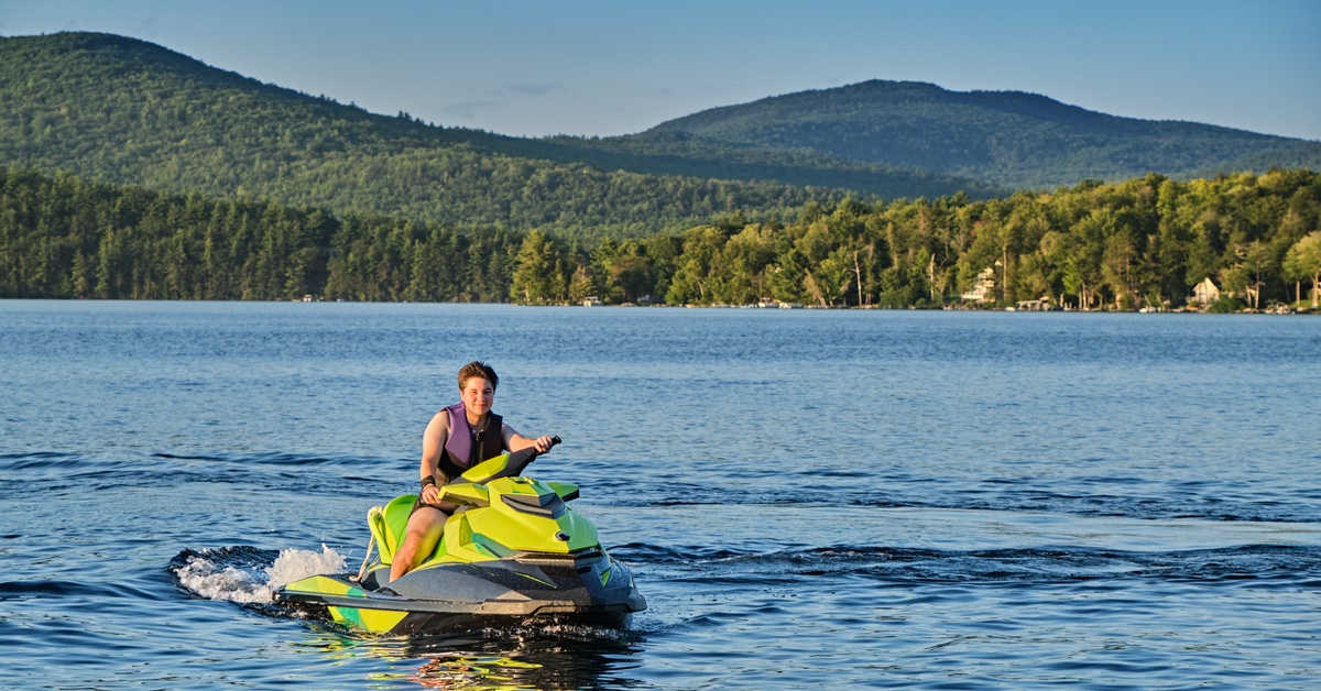 A young person rides a neon yellow jet ski on calm waters. Behind the jet ski is a shoreline full of lush, green trees.