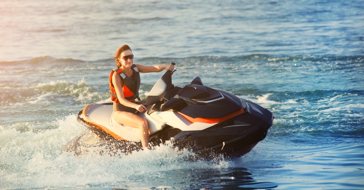 A young adult woman rides a jet ski in a life jacket. She's wearing large sunglasses to protect her face.
