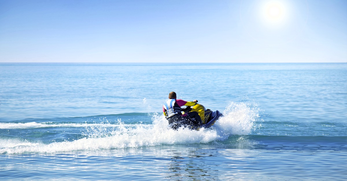 A person riding their jet ski on calm waters and a sunny day. The person is wearing a safety vest and a T-shirt.