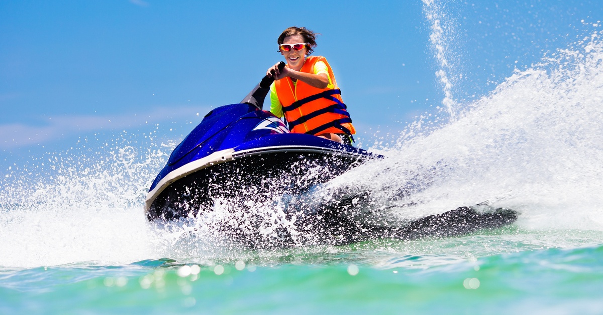 A person wears a bright orange safety vest as they ride a jet ski on the open water. It's a sunny day outside.