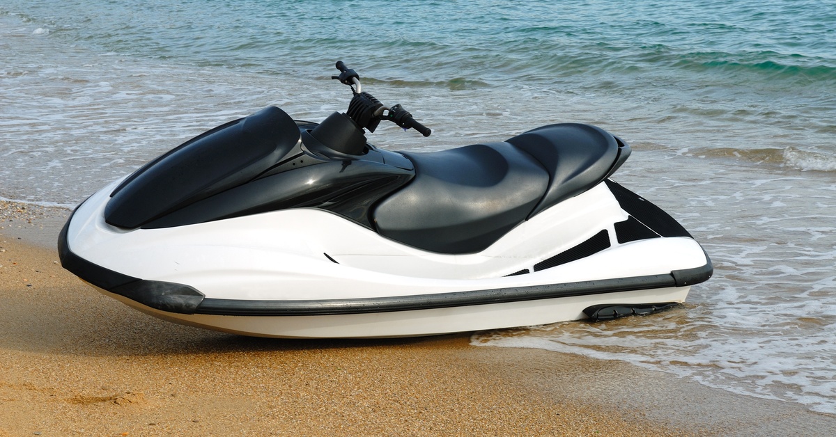 A black and white jet ski sits on the shoreline by itself. The water is calm and it's bright outside.