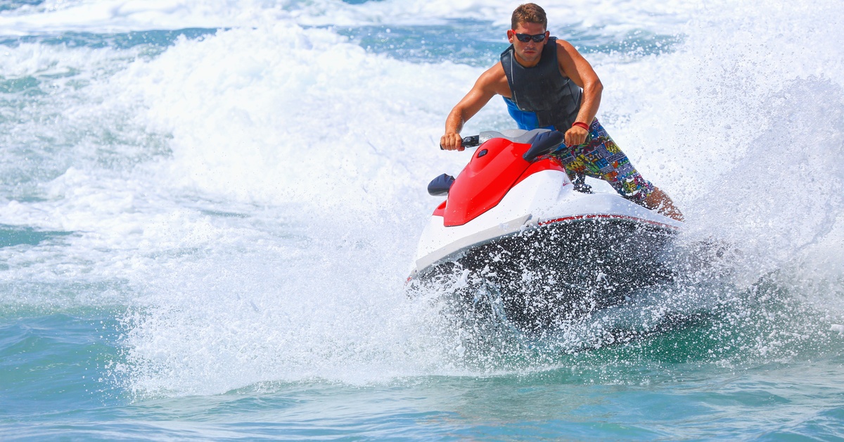 A man rides his red and white jet ski on the water on a sunny day. He wears sunglasses and a safety vest.