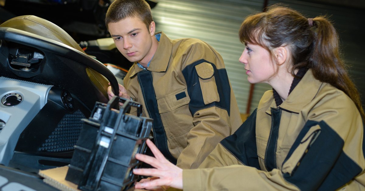 Two jet ski mechanics working on a PWC in the garage. They have the battery out and are wearing tan uniforms.