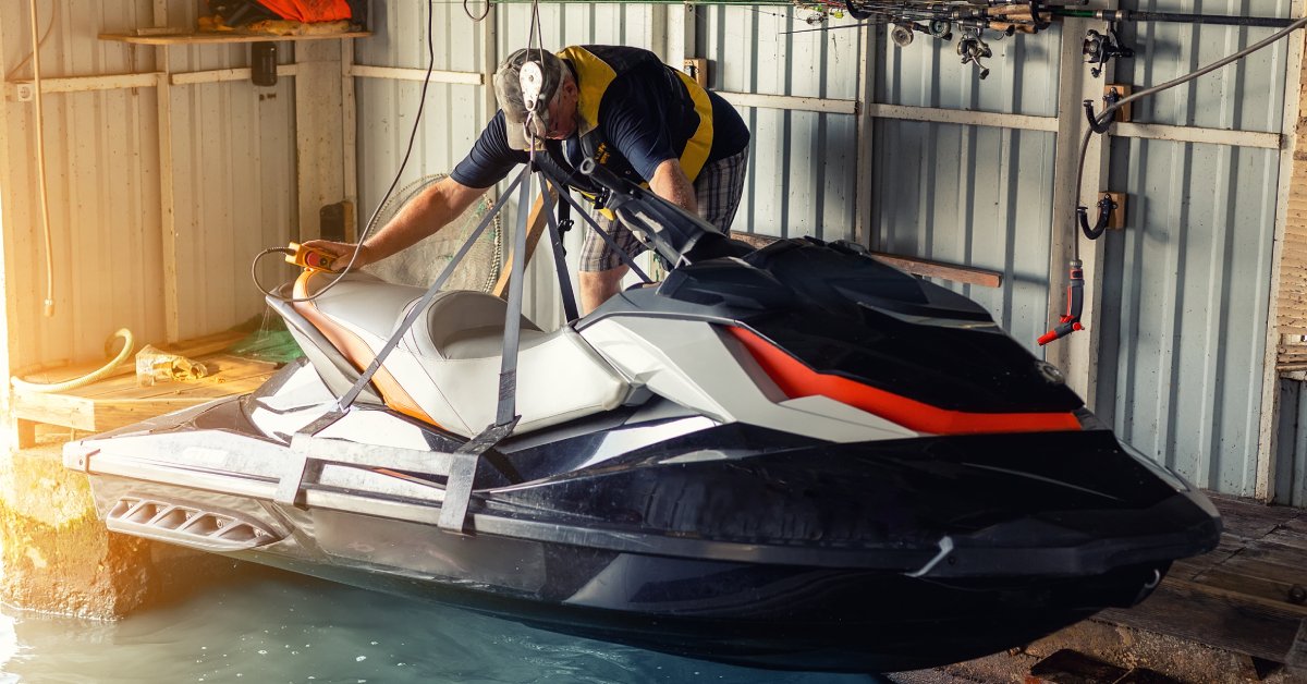 A person stands on the side of a jet ski lift as the black and white PWC lifts up out of the water in hoist straps.