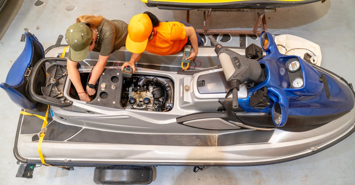 Two mechanics working on a jet ski in the shop. The top of the seat is off, exposing internal components.
