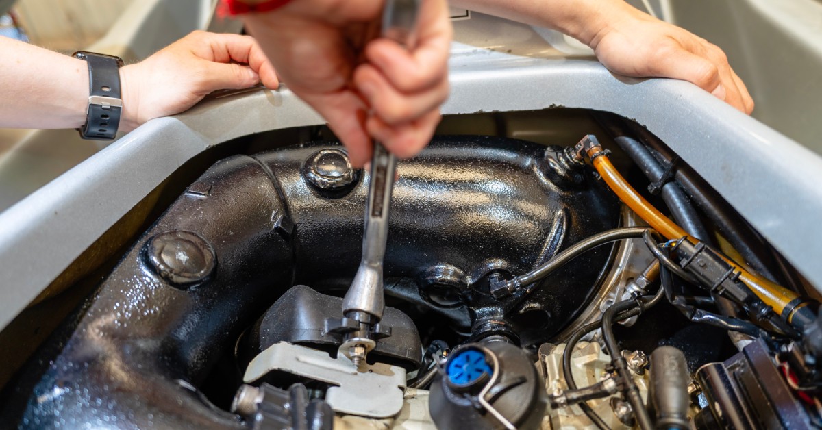 A set of hands works inside the body of a jet ski engine compartment. They hold a silver wrench to disconnect parts.