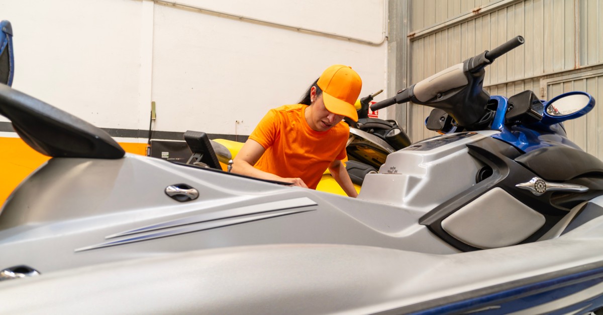 A woman stands in an orange T-shirt and hat next to a jet ski in the garage. The jet ski is silver in color.