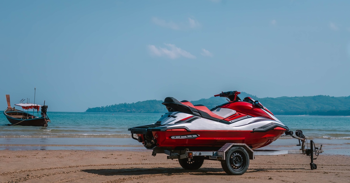 A red and white jet ski sits on a trailer on the beach. The trailer only has two wheels and a hitch in the front.