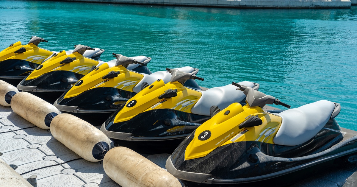 A row of five yellow and black jet skis sit parked at the dock on a sunny day. The water is blue and calm.